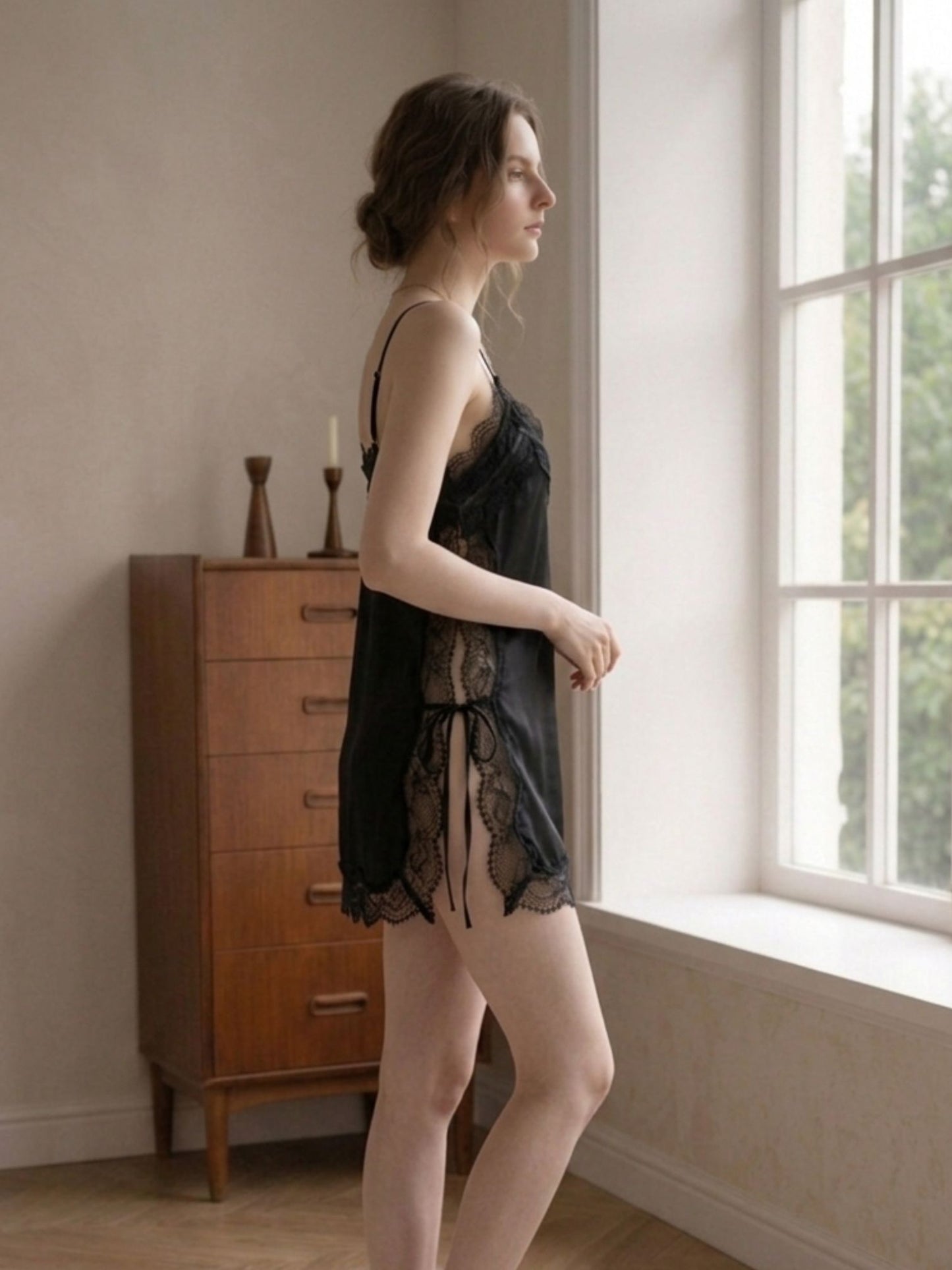 Woman in black lace lingerie standing by a window in a softly lit room.
