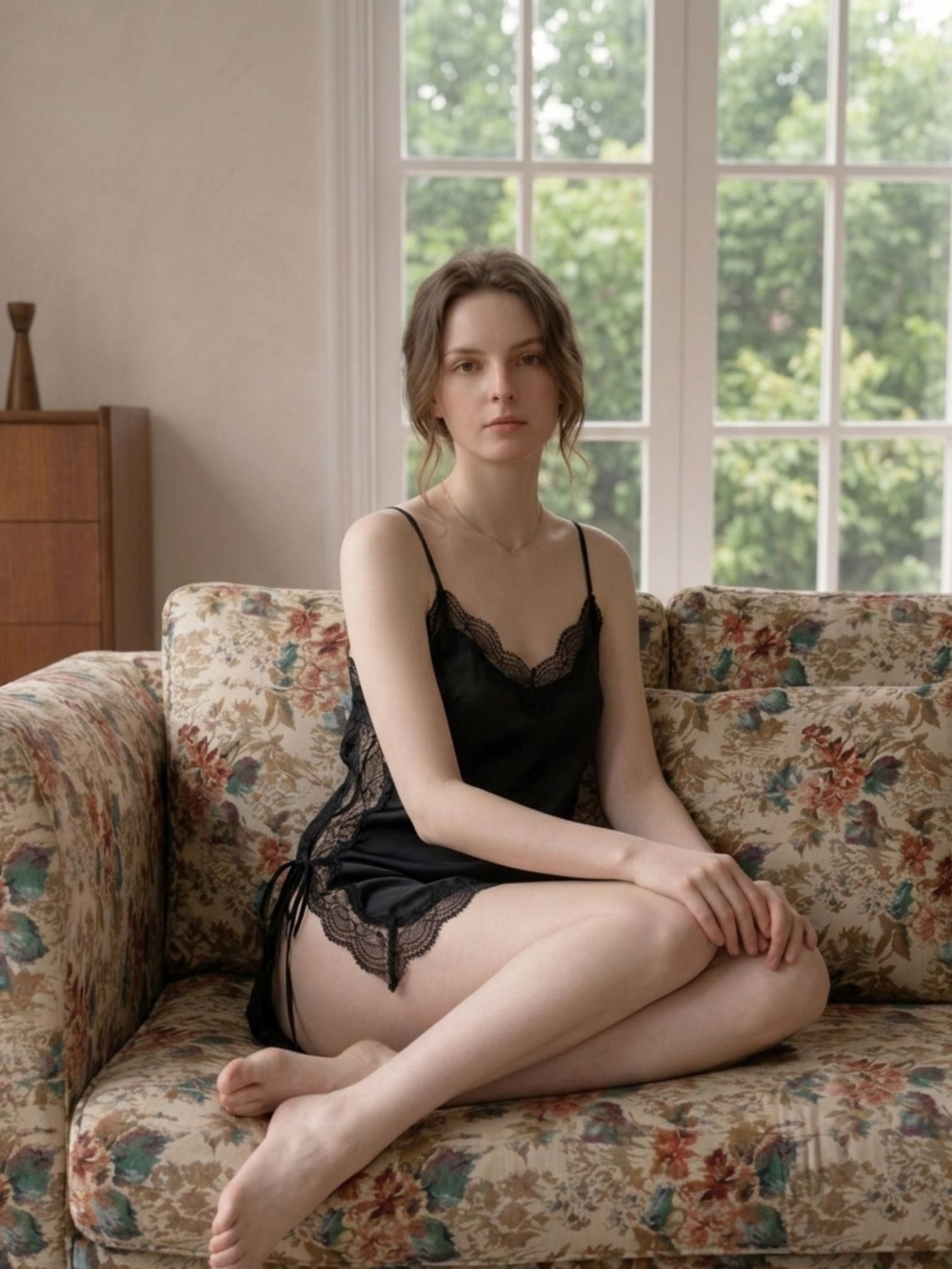 Woman in black lace lingerie sitting on a floral-patterned couch with a window in the background