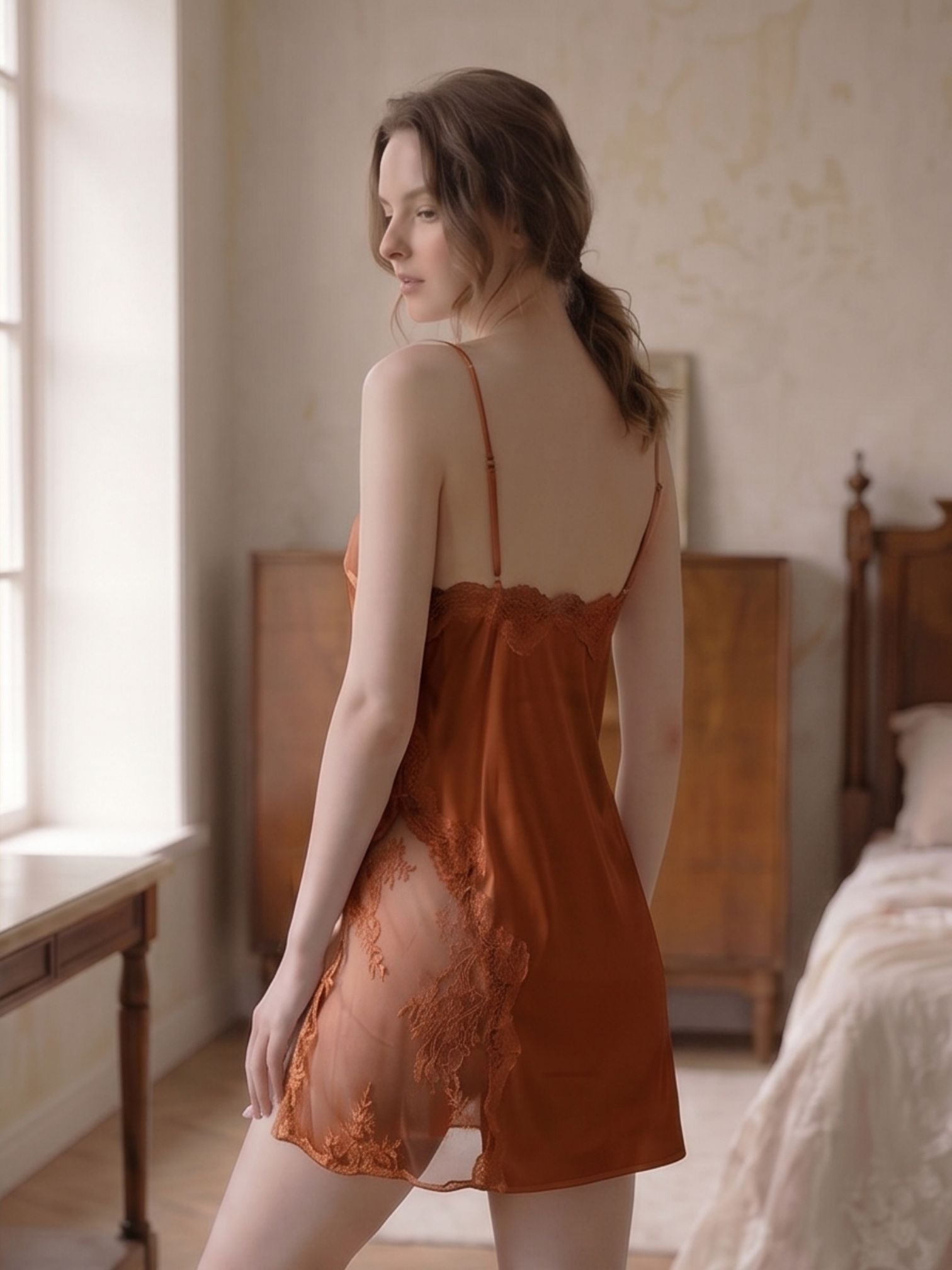 Woman in a rust-colored lace nightgown standing in a softly lit bedroom.