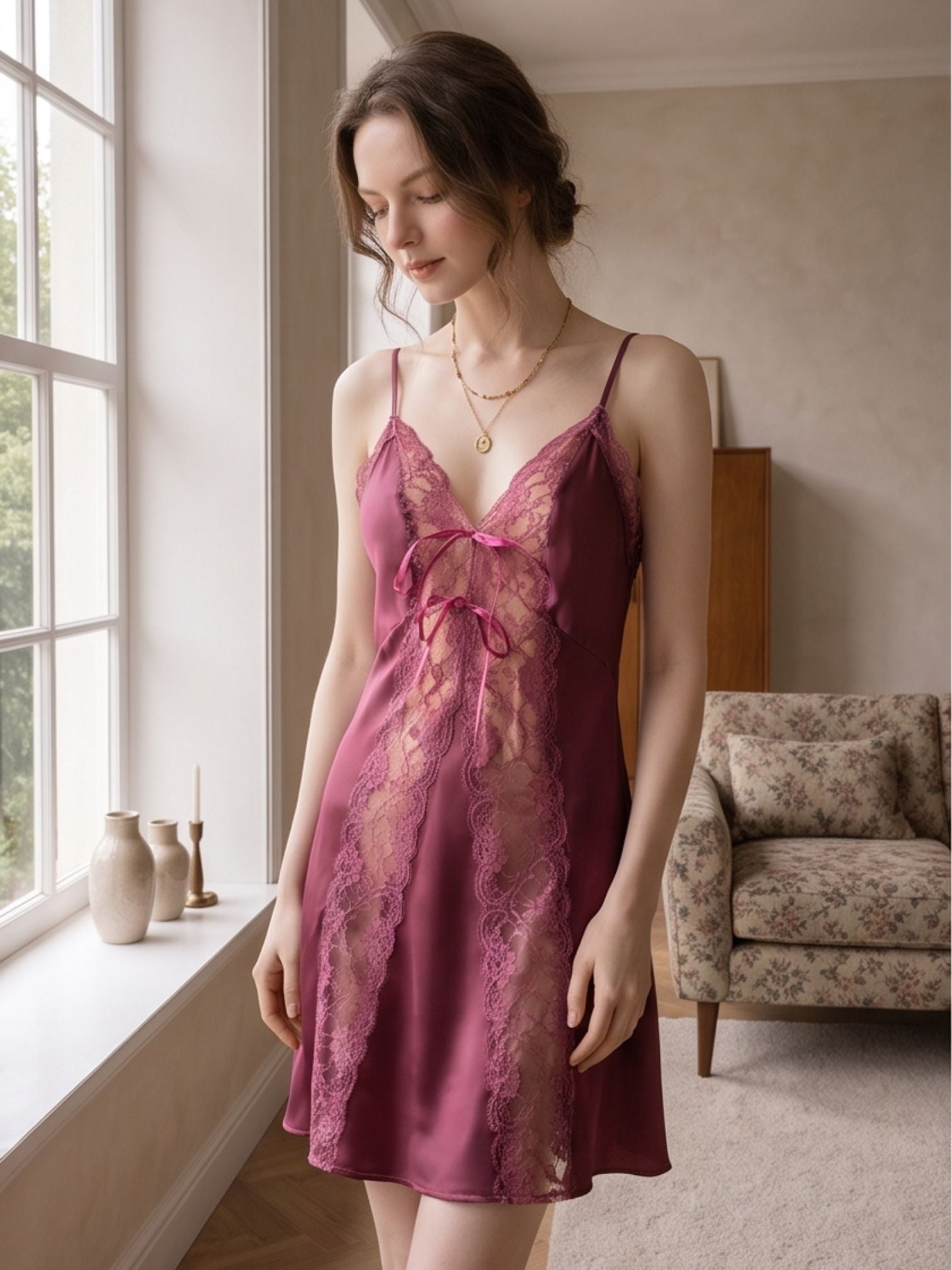 A woman wearing a mauve satin slip dress with pink floral lace panels and a matching lace tie at the bust, standing in a softly lit room by a window.