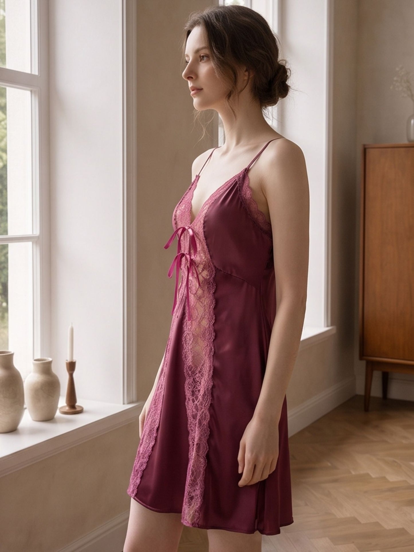A woman wearing a short burgundy satin chemise with lace trim and a front tie, standing in a softly lit room by a window.