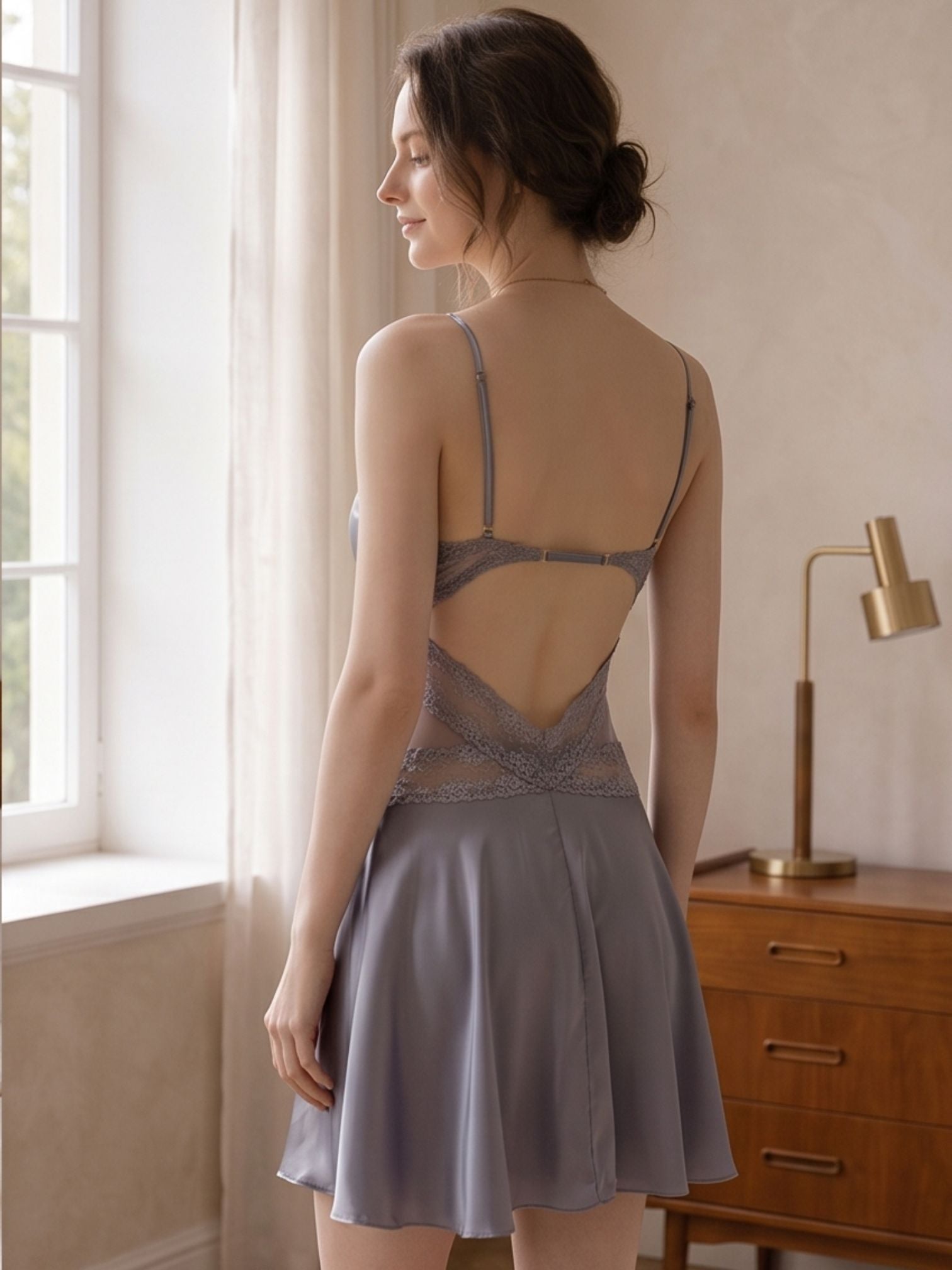 A woman stands in a softly lit bedroom wearing a short satin slip dress with thin straps, a lace-trimmed open back, and a flared skirt. She is shown from the back near a window, with a wooden dresser and lamp in the background.