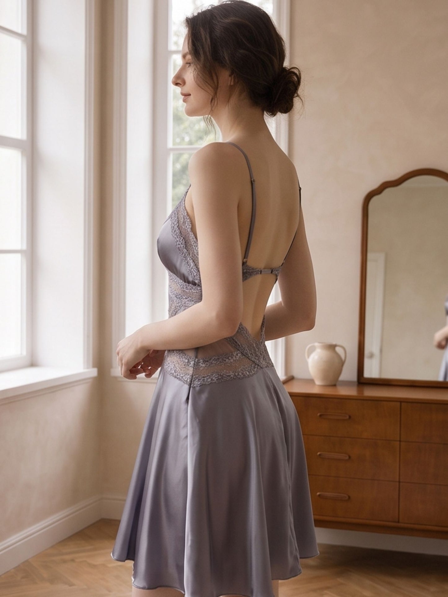 A woman stands by a window in a softly lit room wearing a sleeveless, backless sleepwear dress in a muted lavender-gray color with lace detailing.