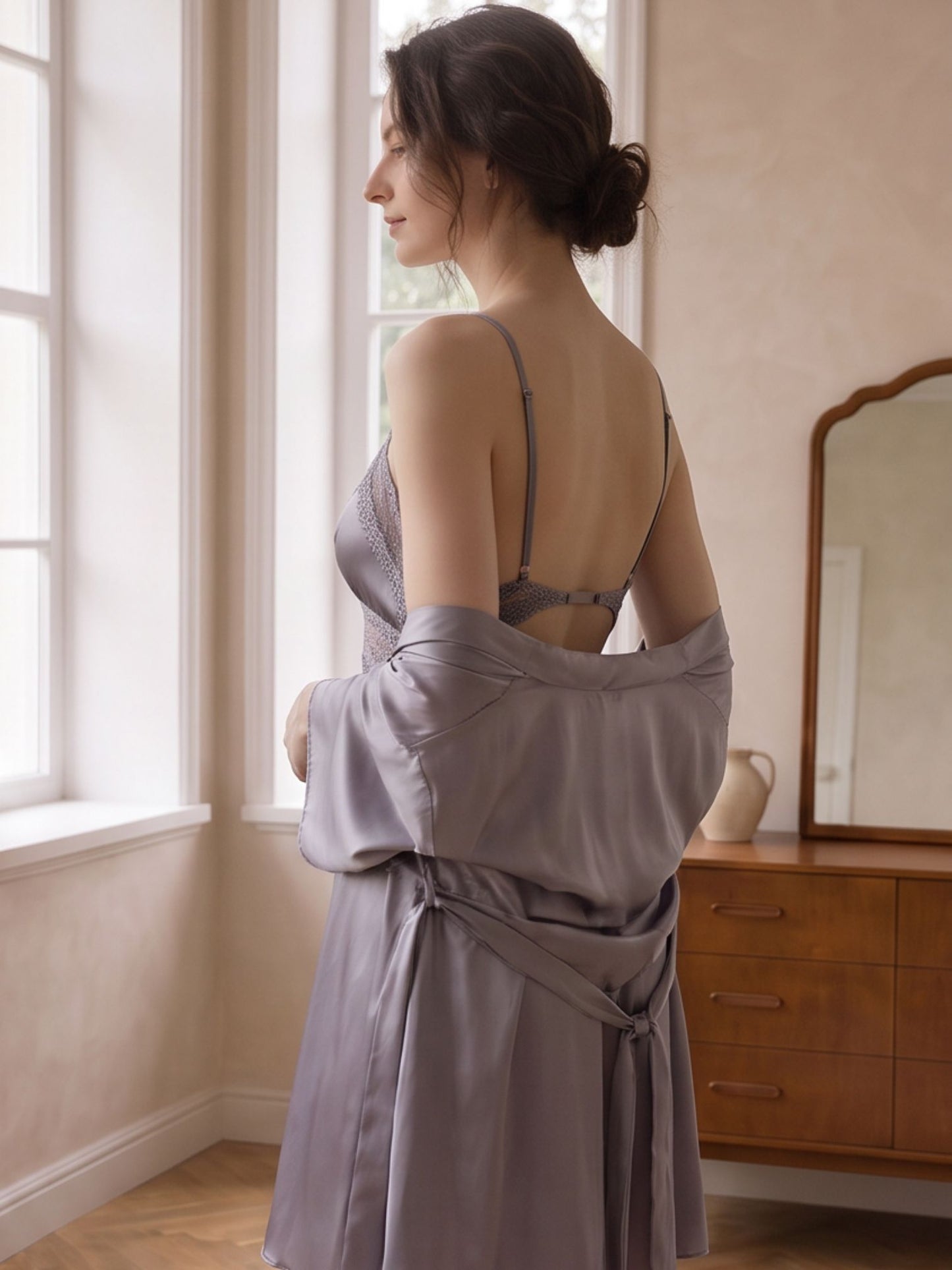 A woman stands by a window in a softly lit room, wearing a lavender-gray satin robe over a matching lace lingerie set. The robe is tied at the waist and draped off her shoulders, with a wooden dresser and mirror visible in the background.