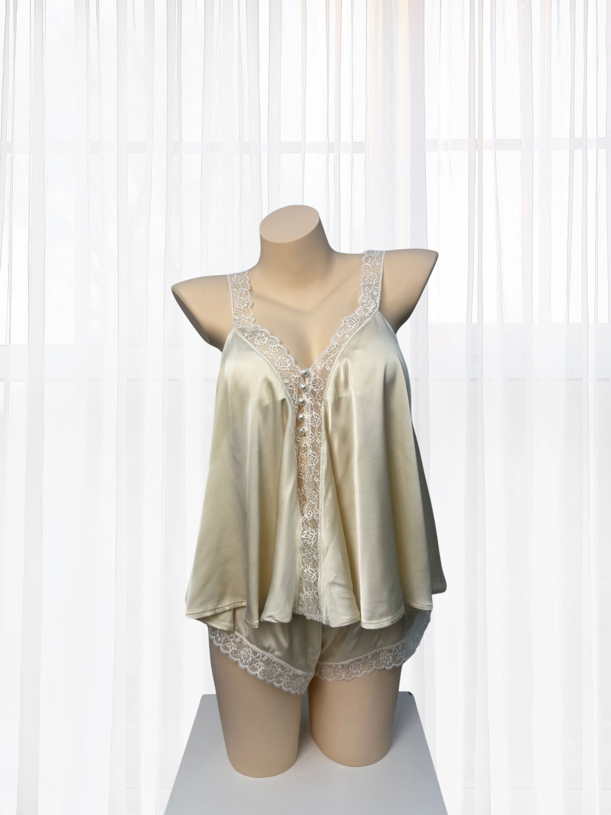 A two-piece satin pajama set displayed on a mannequin torso, featuring a camisole top and matching shorts in a light champagne beige color with white lace trim, set against a background of white sheer curtains.