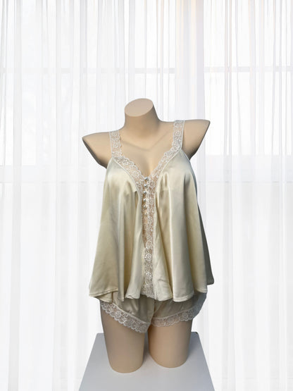 A two-piece satin pajama set displayed on a mannequin torso, featuring a camisole top and matching shorts in a light champagne beige color with white lace trim, set against a background of white sheer curtains.
