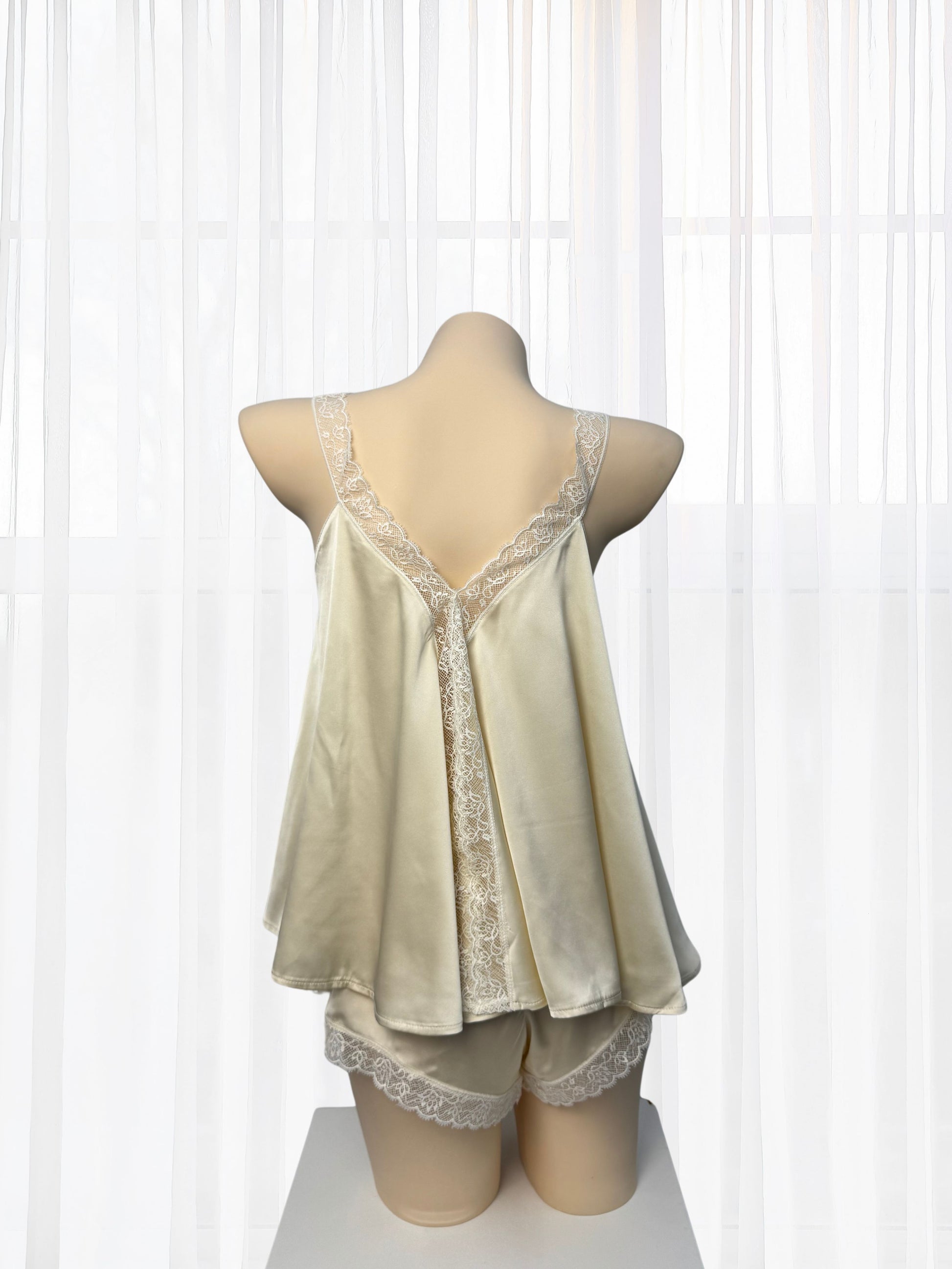 A cream-colored satin pajama set with lace trim displayed on a mannequin torso against a white curtain backdrop.